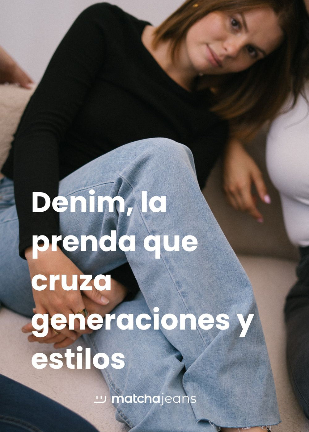Chica con pelo corto pelirrojo está sentada en un sofá con las piernas cruzadas, lleva una camiseta de manga larga negra y vaqueros azules